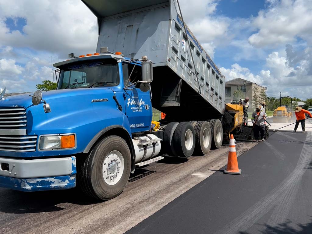 Paving company near me in Fort Lauderdale 3-D Paving drop a fresh truckload of hot asphalt into a paving machine laying down a final lift of asphalt on a residential roadway.