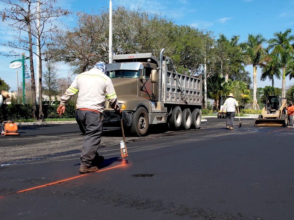 Asphalt Paving crew member marks out lines with spray paint defining where the next pull of asphalt for an asphalt overlay will be installed. Brown dump truck in the background near a skid steer. New asphalt parking lot by 3-D Paving and Sealcoating, South Florida's Commercial Paving Experts. Learn from the American Asphalt Paving experts at 3-D Paving and Sealcoating in Coral Springs, FL.