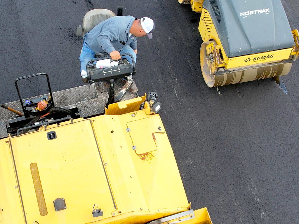 Aerial view of a construction worker on an asphalt paving machine to lay a bottom lift of hot mix asphalt with an asphalt rolling machine in the background. 3-D Paving